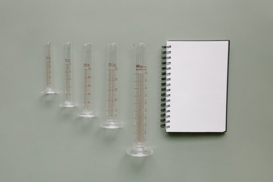 A collection of graduated cylinders next to a spiral notebook on a green background.