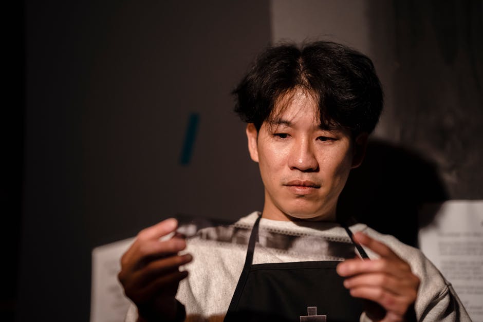 Asian man inspecting a vintage photographic film strip in a darkroom setting.