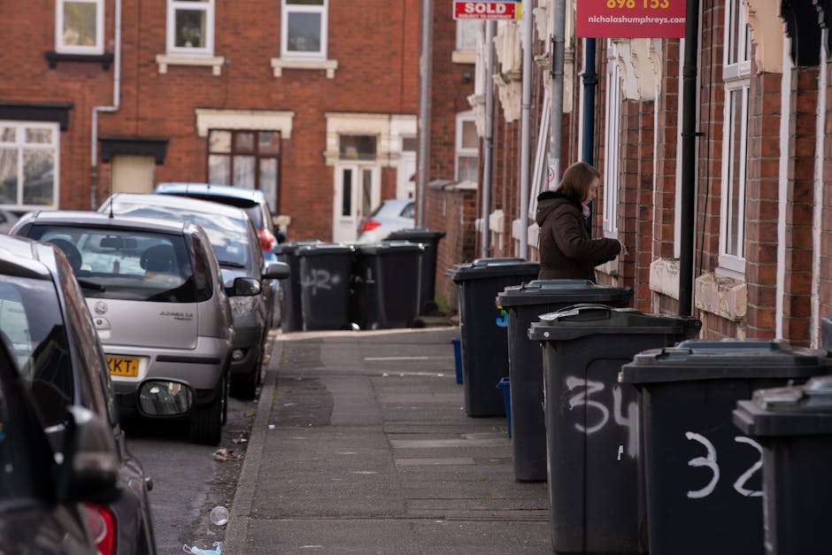 A street scene in a residential area with garbage bins and parked cars along the sidewalk.