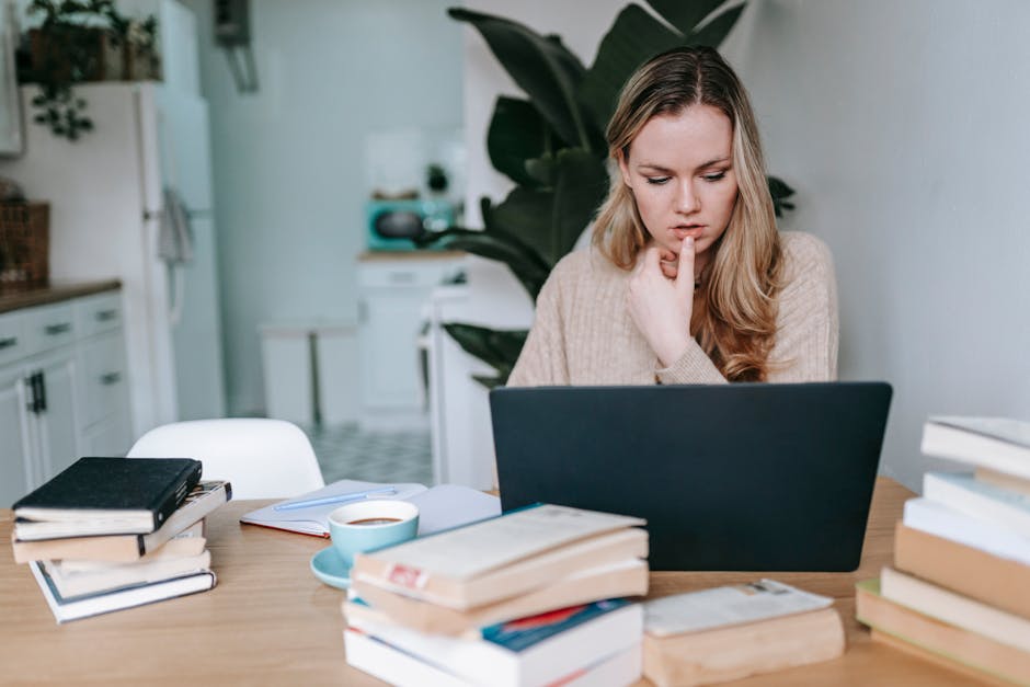 Serious intelligent female studying online on netbook at table with cup of coffee and textbooks on blurred background of home