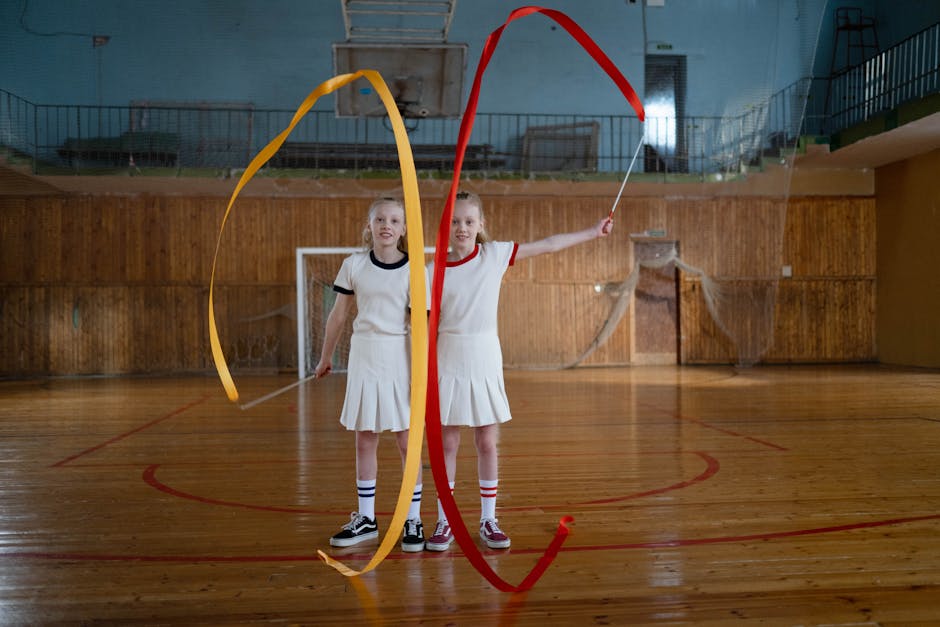 Two young girls performing rhythmic gymnastics with ribbons in an indoor sports hall.