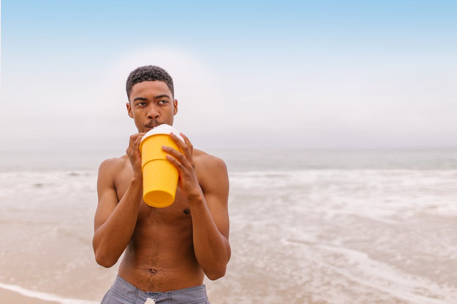 A shirtless man drinking from a yellow cup at the beach, enjoying a sunny seaside day.