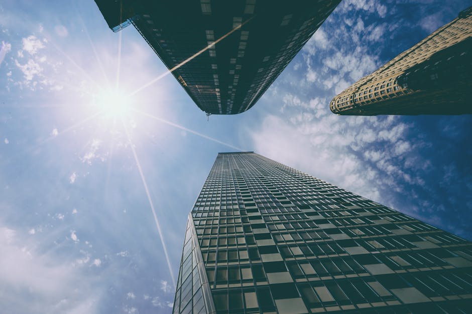 Low angle view of tall skyscrapers with sun glare against a bright blue sky.