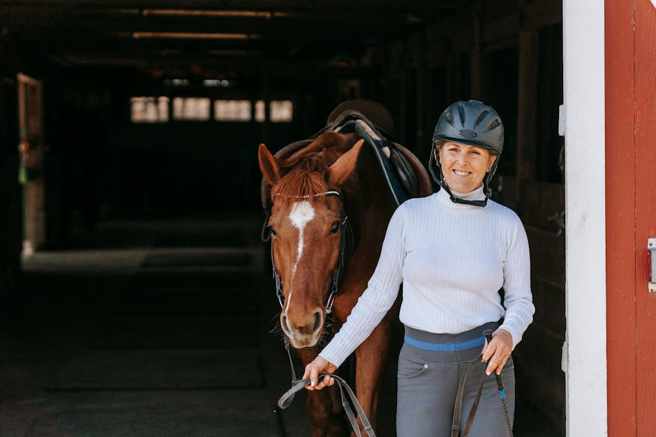 A smiling woman in riding gear leading a saddled horse outdoors.