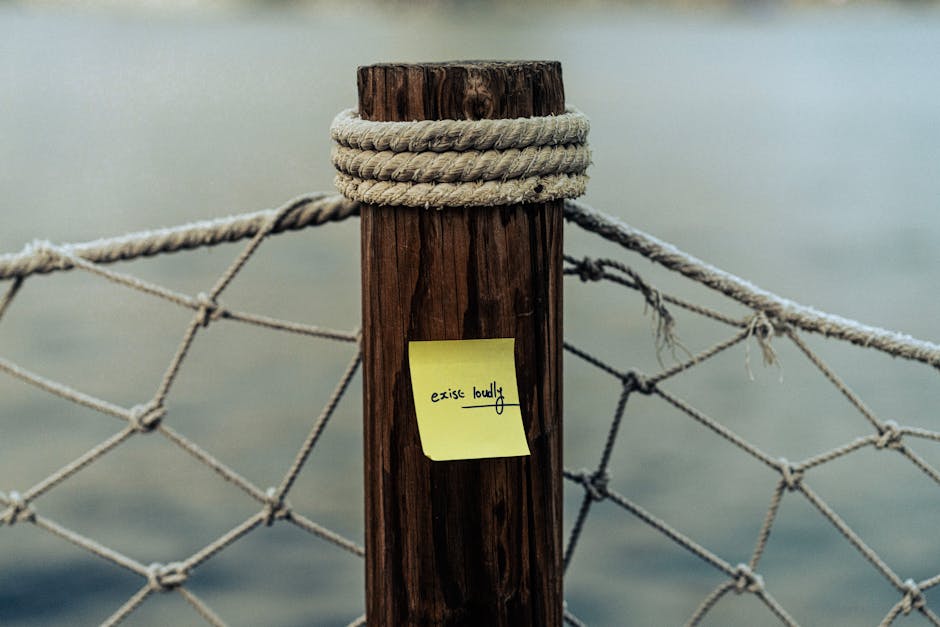 Close-up of a sticky note with 'exist loudly' on a rope-wrapped post by water.