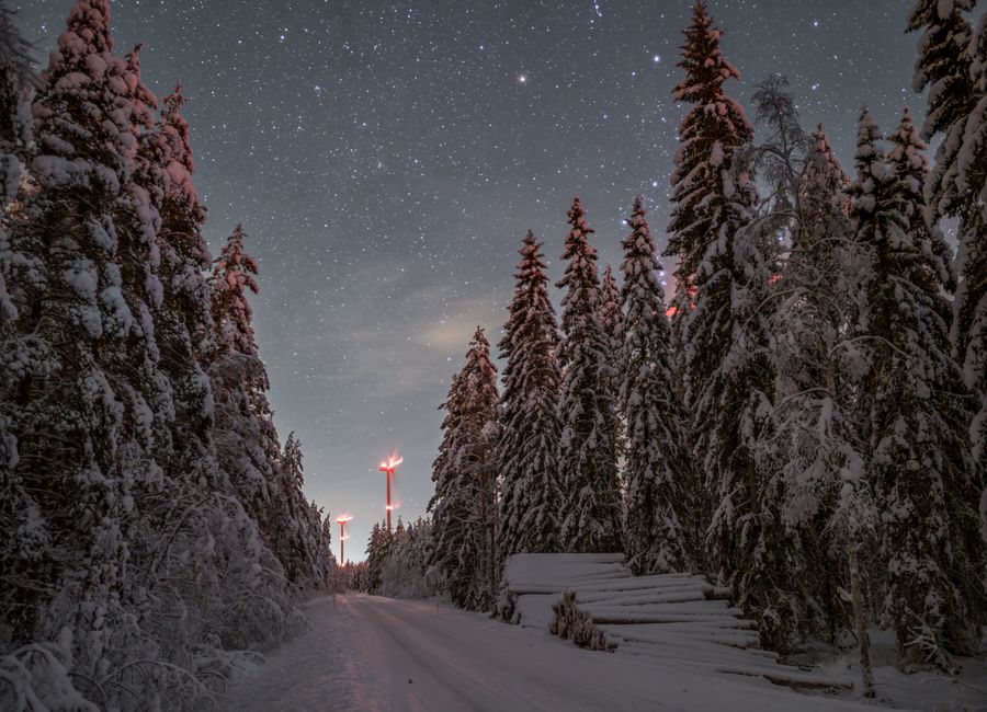 Scenic winter night with starry sky over snow-covered forest road and wind turbines in the distance.