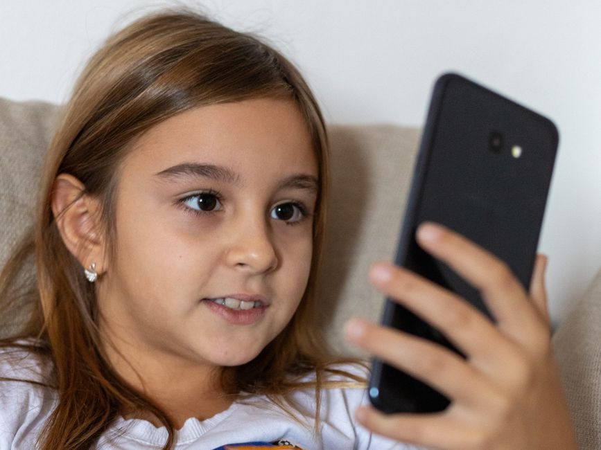 A young girl with long brown hair smiles while using a smartphone indoors.