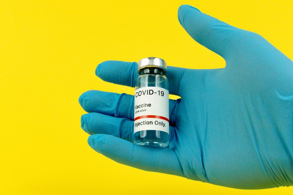 A gloved hand holds a COVID-19 vaccine vial against a bright yellow background.
