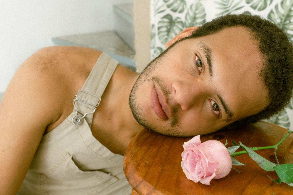 A contemplative man with a beard leans on a wooden surface beside a pink rose indoors.