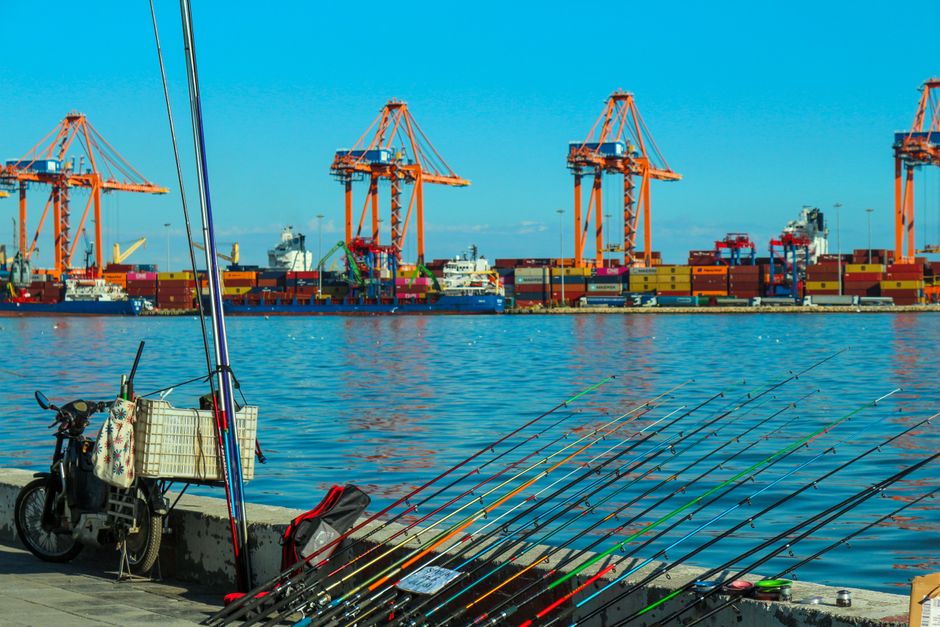 Colorful port view featuring fishing rods, cranes, and shipping containers under a bright blue sky.